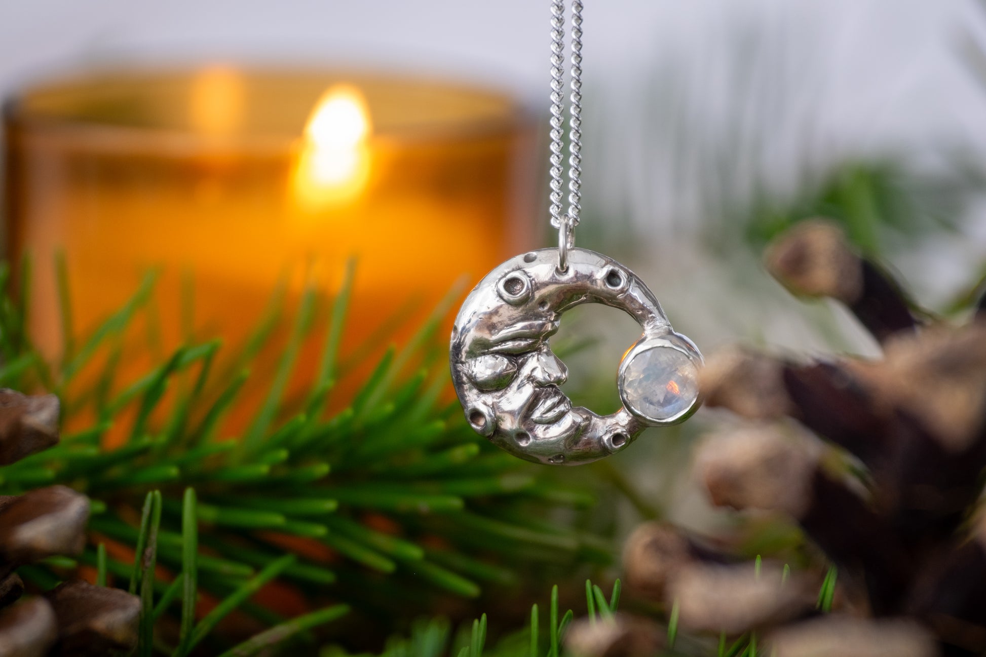 a festive photo of a moon shaped pendant surrounded by pine needles, pine cones and a candle